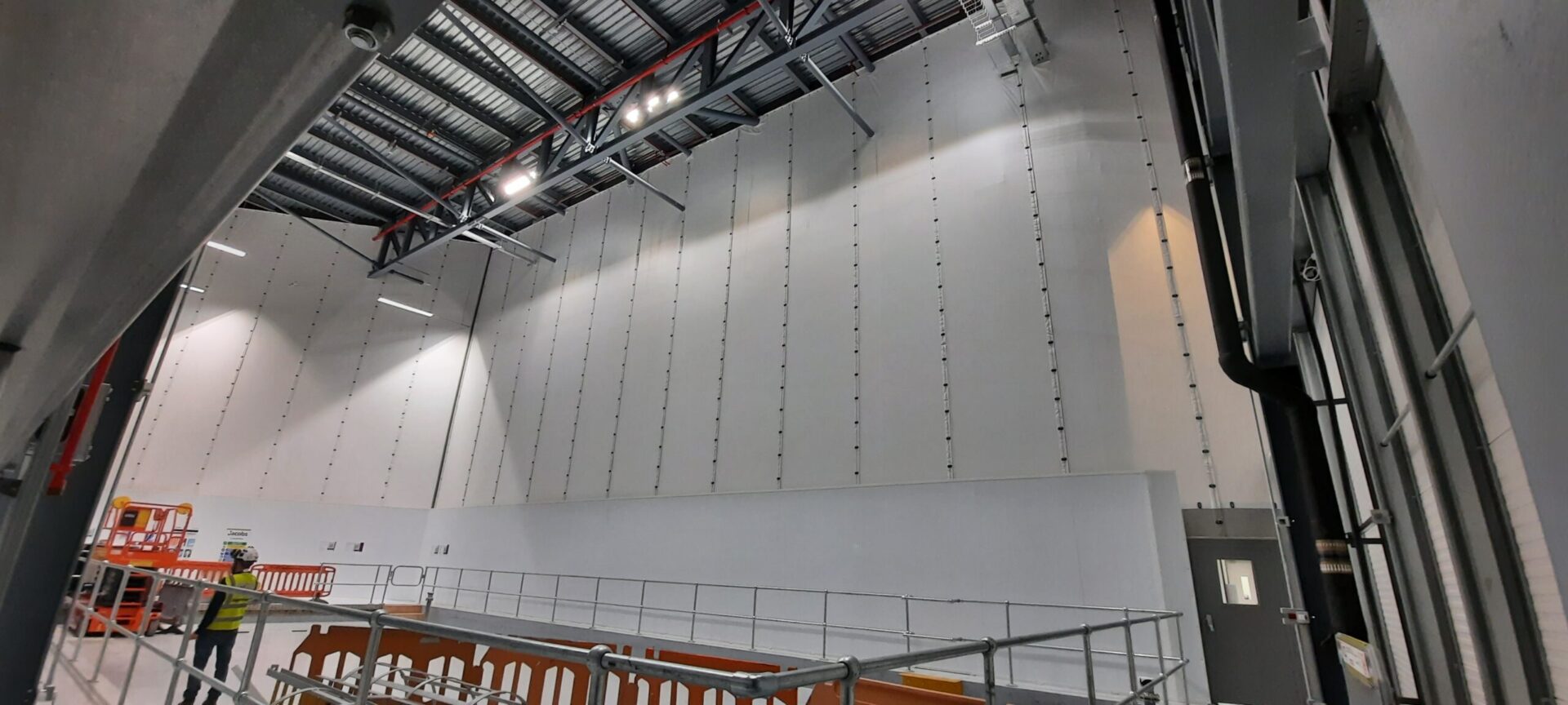 Großflächiges „Flexiscreen“-Industrie-Vorhangfassadensystem in einer pharmazeutischen Produktionsstätte, bei dem raumhohe weiße PVC-Trennwände an Stahldachbindern mit vertikalen Führungsschienen aufgehängt sind und so eine Trennwand in Reinraumqualität bilden. Im Vordergrund ist ein Mitarbeiter in gut sichtbarer Schutzausrüstung und mit Schutzhelm zu sehen, neben provisorischen Gerüstabsperrungen und einer orangefarbenen Scherenhebebühne, während industrielle LED-Beleuchtung den Innenraum des Hochregallagers ausleuchtet. 