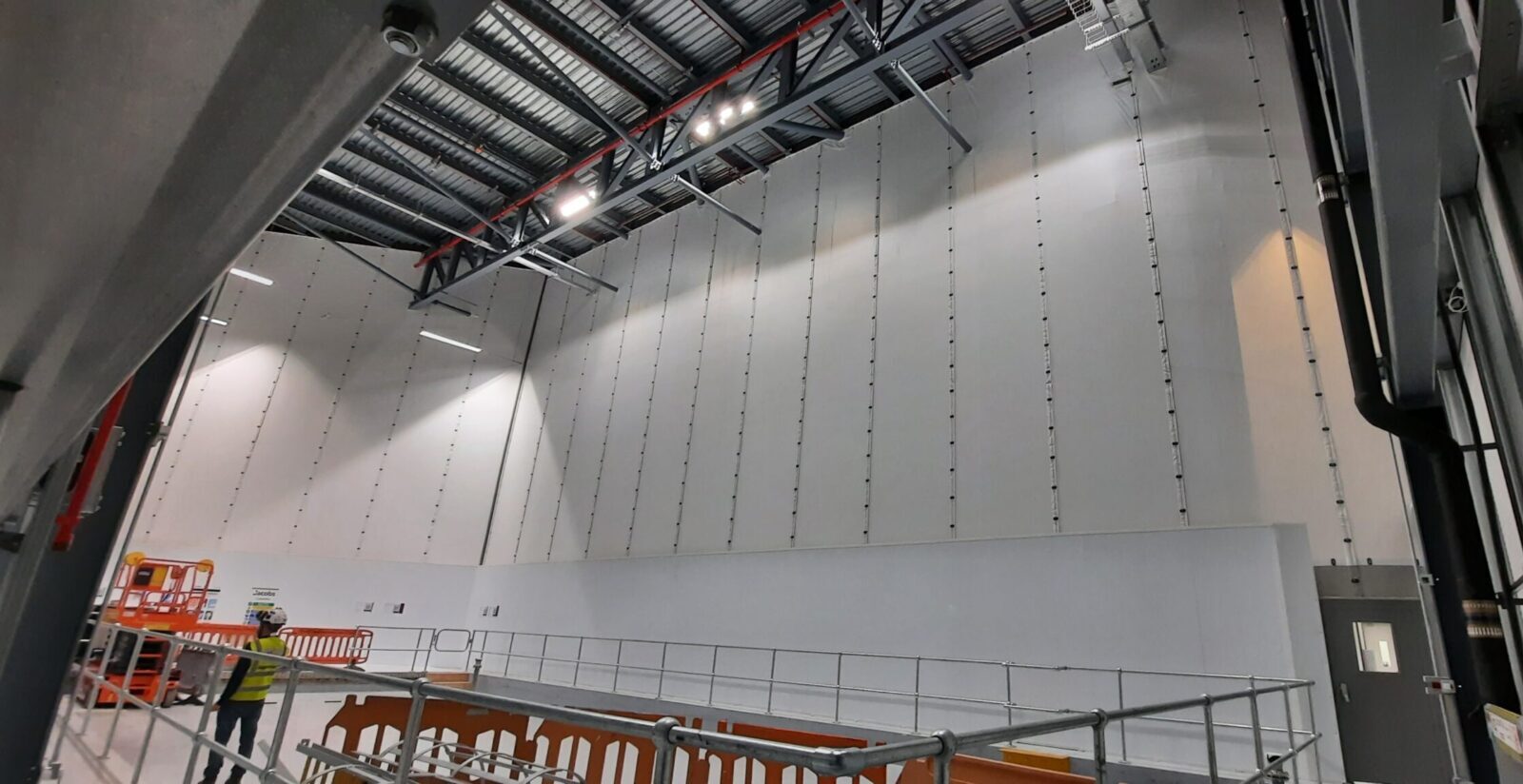 Großflächiges „Flexiscreen“-Industrie-Vorhangfassadensystem in einer pharmazeutischen Produktionsstätte, bei dem raumhohe weiße PVC-Trennwände an Stahldachbindern mit vertikalen Führungsschienen aufgehängt sind und so eine Trennwand in Reinraumqualität bilden. Im Vordergrund ist ein Mitarbeiter in gut sichtbarer Schutzausrüstung und mit Schutzhelm zu sehen, neben provisorischen Gerüstabsperrungen und einer orangefarbenen Scherenhebebühne, während industrielle LED-Beleuchtung den Innenraum des Hochregallagers ausleuchtet. 